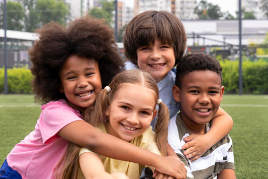 A diverse group of four smiling children hugging each other and posing outdoors on a grassy field. The children are enjoying their time together, showcasing friendship and happiness