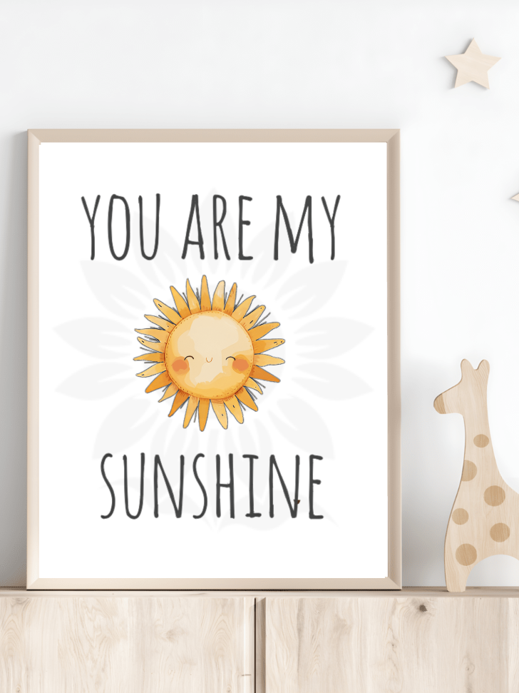 Nursery room decor with framed wall art of a cheerful, smiling sun and the text 'YOU ARE MY SUNSHINE.' The design, from Blossoming Minds, is displayed on a shelf next to a wooden giraffe toy, encouraging positive affirmations for children.